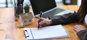 Woman's hands writing on paper with miniature house on desk